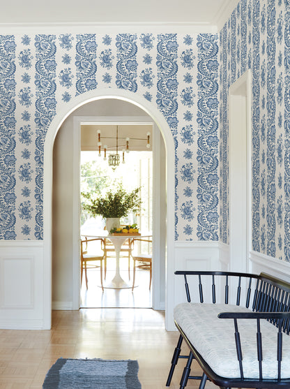 Blue and white damask wallpaper in a white wainscoted foyer; arch doorway to a sunlit dining room with pedestal table.