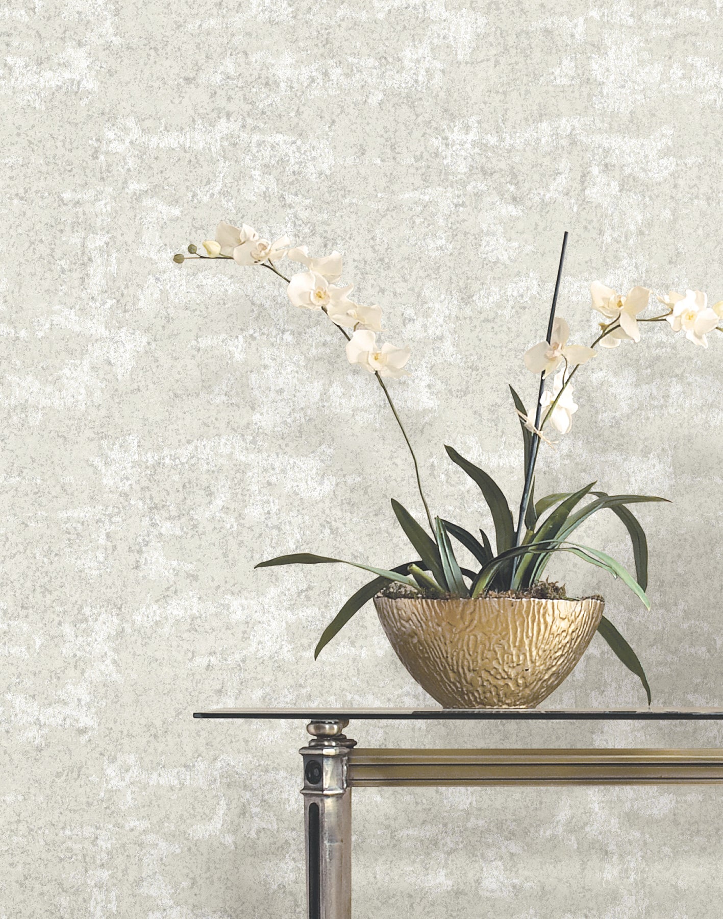 Gold hammered bowl with white orchid stems and green leaves on a glass-top console table against a textured gray wall.