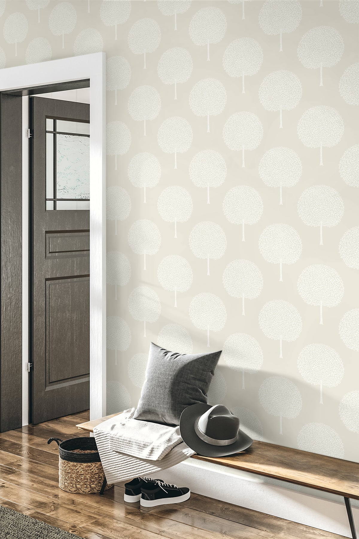 Neutral entryway with gray tree-pattern wallpaper, white door frame, wooden bench, grey cushion, hat, magazines, sneakers.