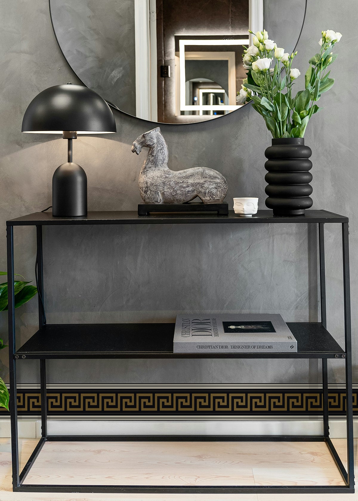 Black metal console with two shelves, lamp, ceramic horse, ribbed black vase with white flowers, round wall mirror.