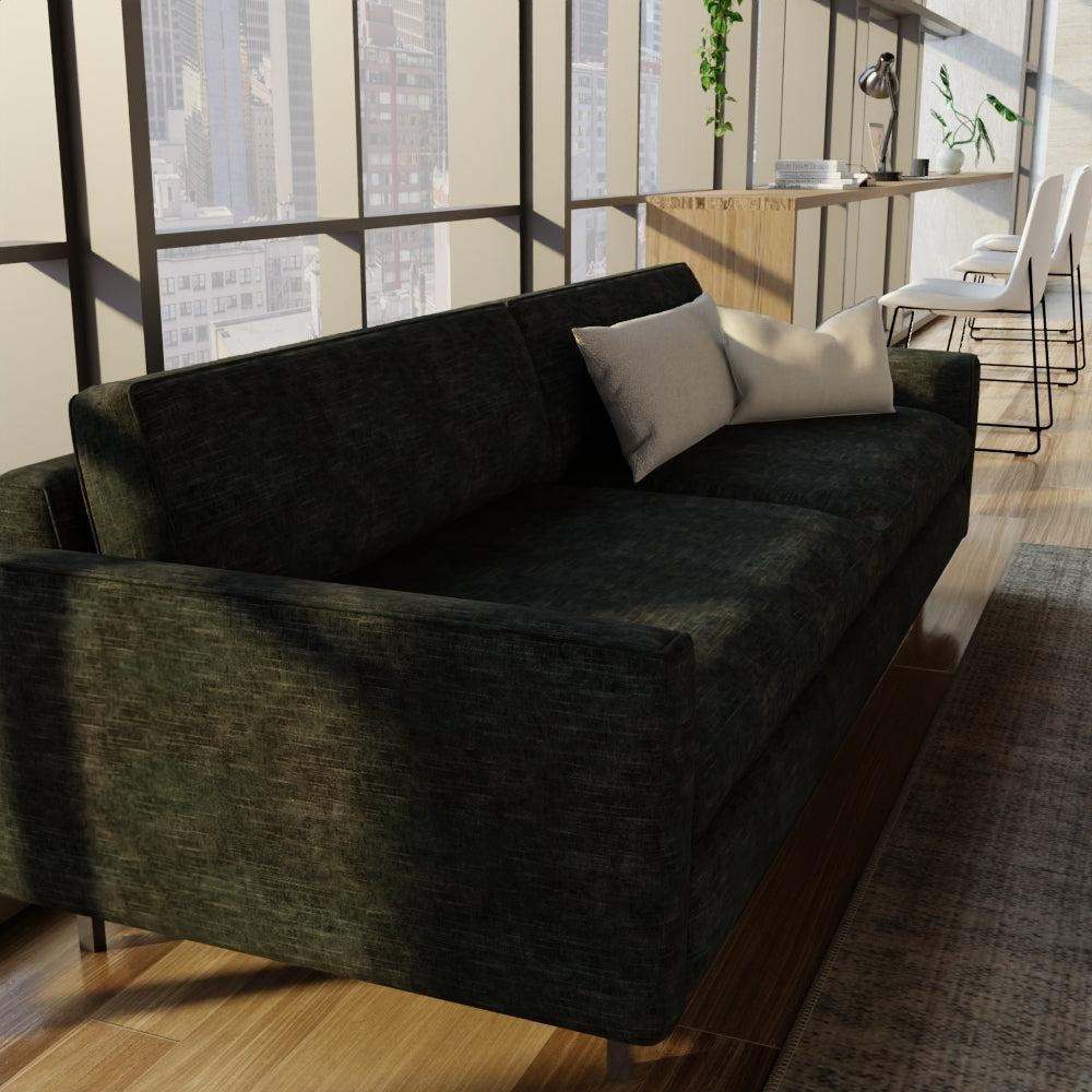 Dark emerald green velvet upholstery on a sofa with boxy arms and two light gray throw pillows in a modern open-plan living space.