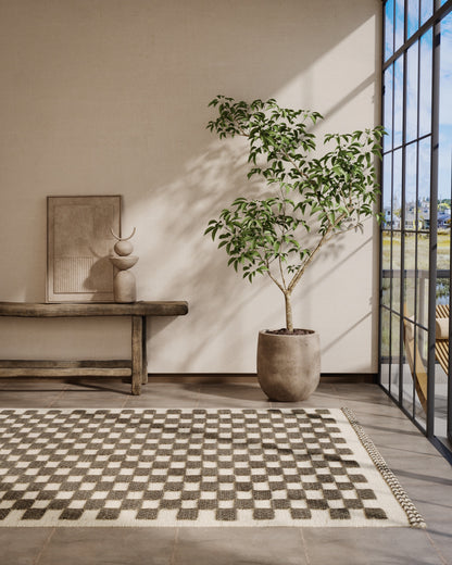 Sunlit beige-walled room with a black-&-white checkered rug, wooden bench, framed art, & a tall leafy tree in a terracotta pot by glass doors.