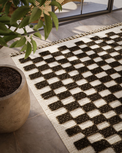 Woven checkered rug with dark brown and cream squares, jute fringe edge, indoor mat by glass doors.