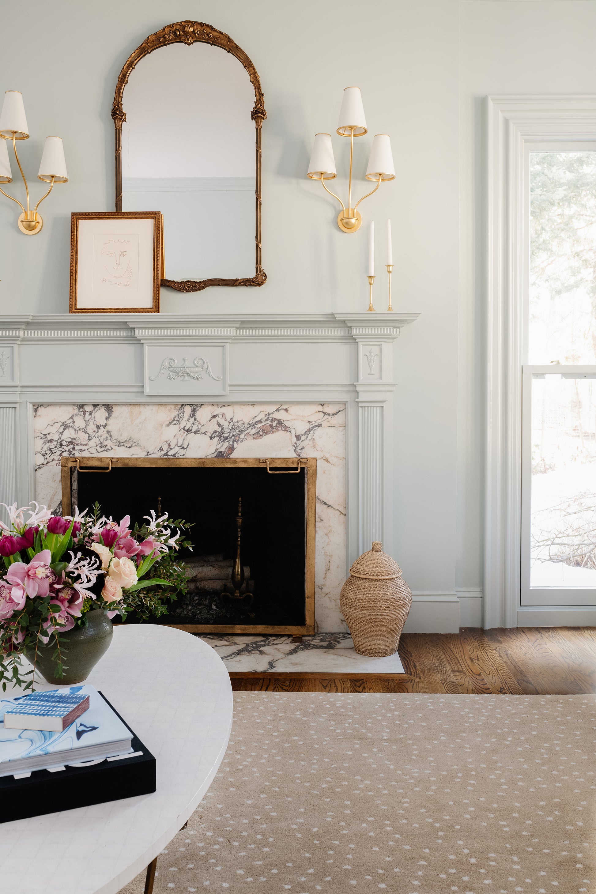 Traditional living room with a rug (2' x 3') on the floor, marble fireplace with gold-trimmed mantel, arch gilt mirror, twin sconces, & flowers on a table.