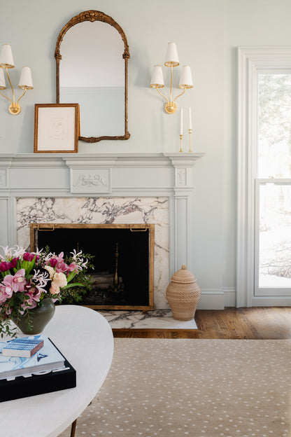 Traditional living room with a rug (2' x 3') on the floor, marble fireplace with gold-trimmed mantel, arch gilt mirror, twin sconces, & flowers on a table.