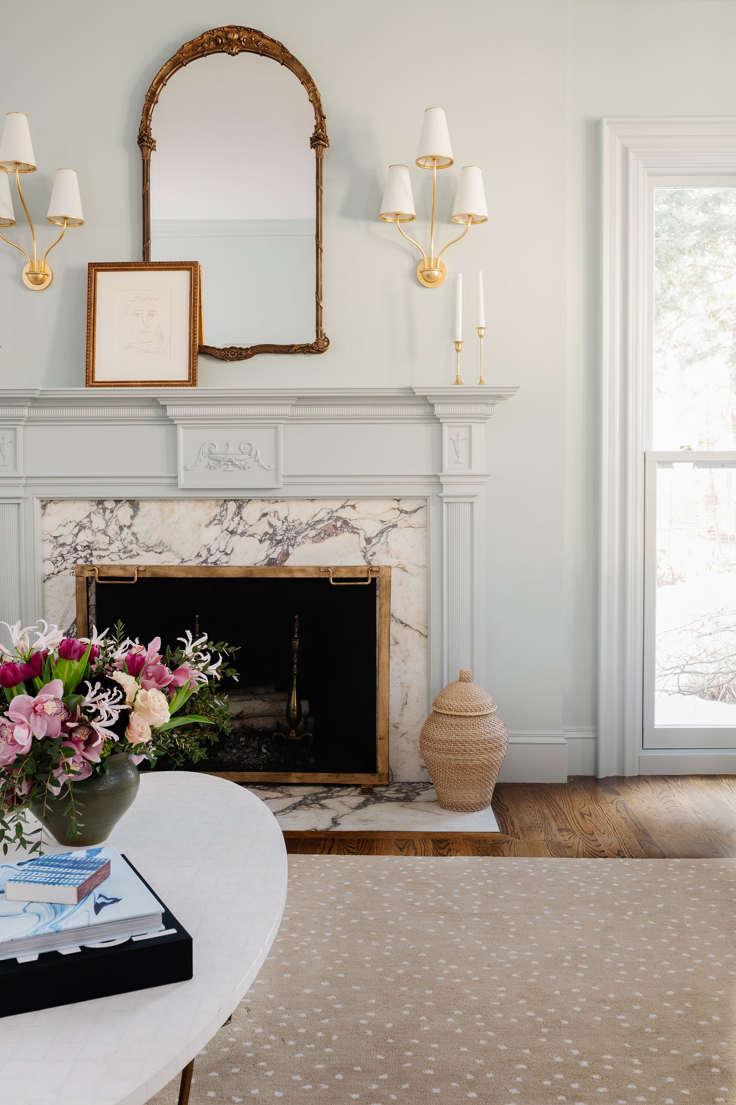 Pastel blue 10' x 14' rug on a living room floor, with a marble fireplace, gold arched mirror, sconces, framed art, wicker basket, and flowers.