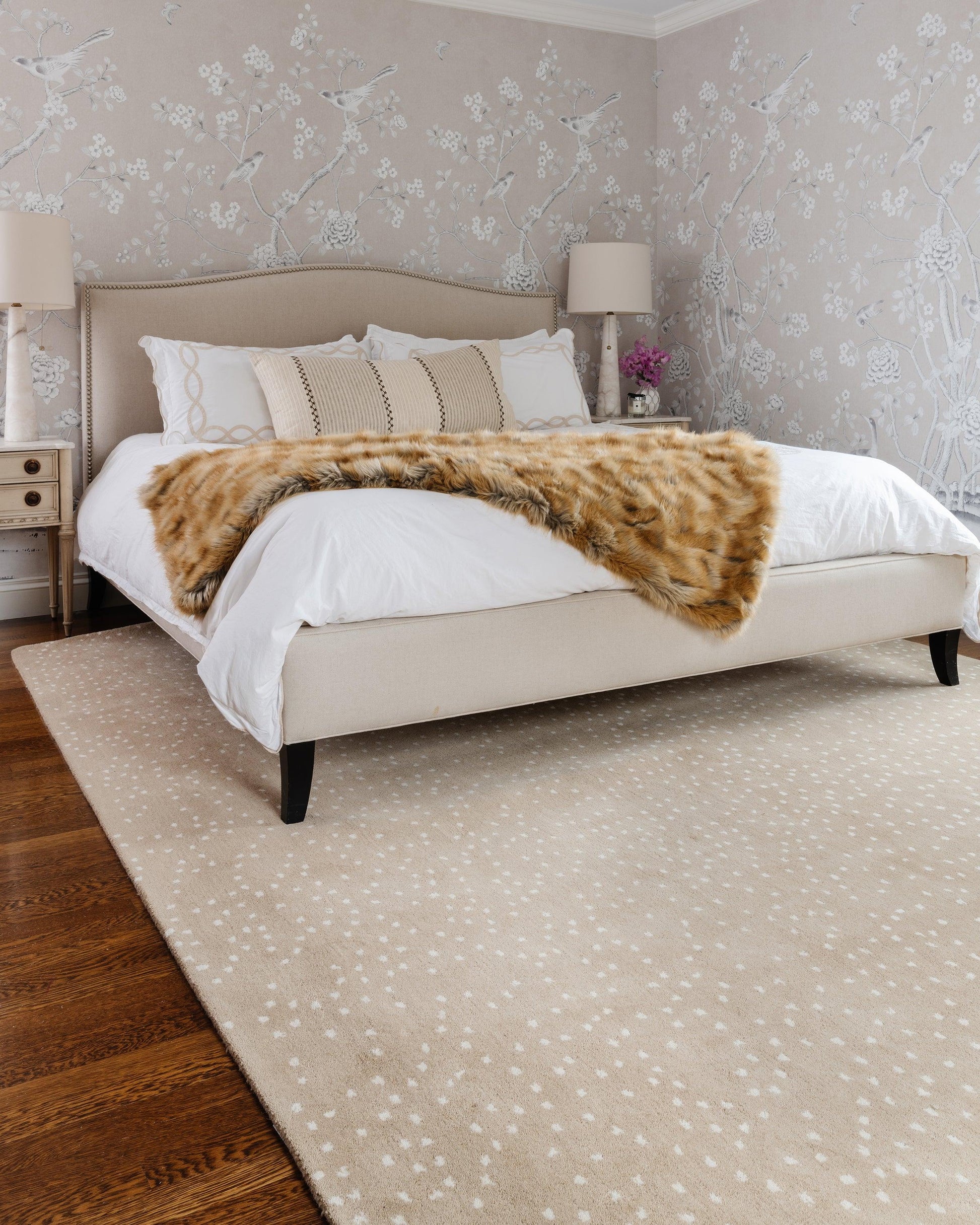 Beige 2' x 3' rug with a light dotted pattern, shown on a light floor with wooden nightstands and floral wallpaper in the background.