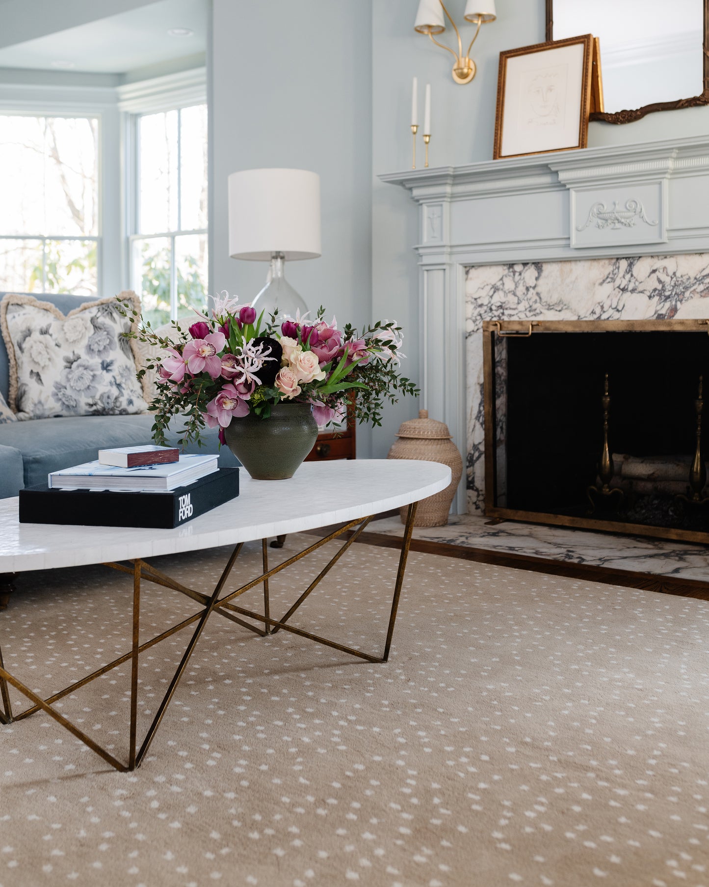 Rug in a light blue living room on the floor, with an oval white coffee table on gold geometric legs, pink floral arrangement, lamp, and marble fireplace.