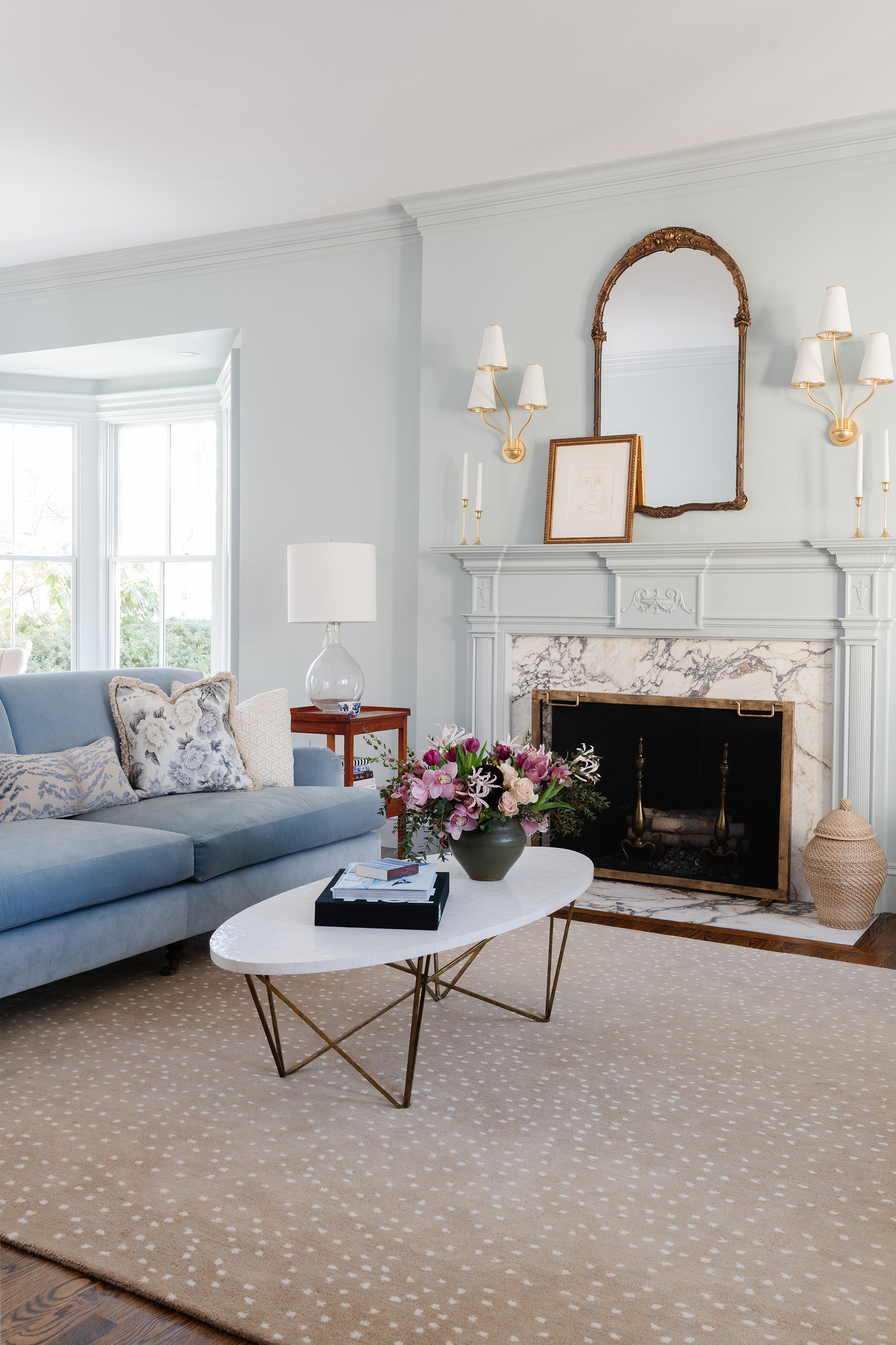 Soft blue rug in a living room with a marble fireplace, gold-framed mirror, white oval coffee table, and a floral bouquet; rug 2' x 3'.