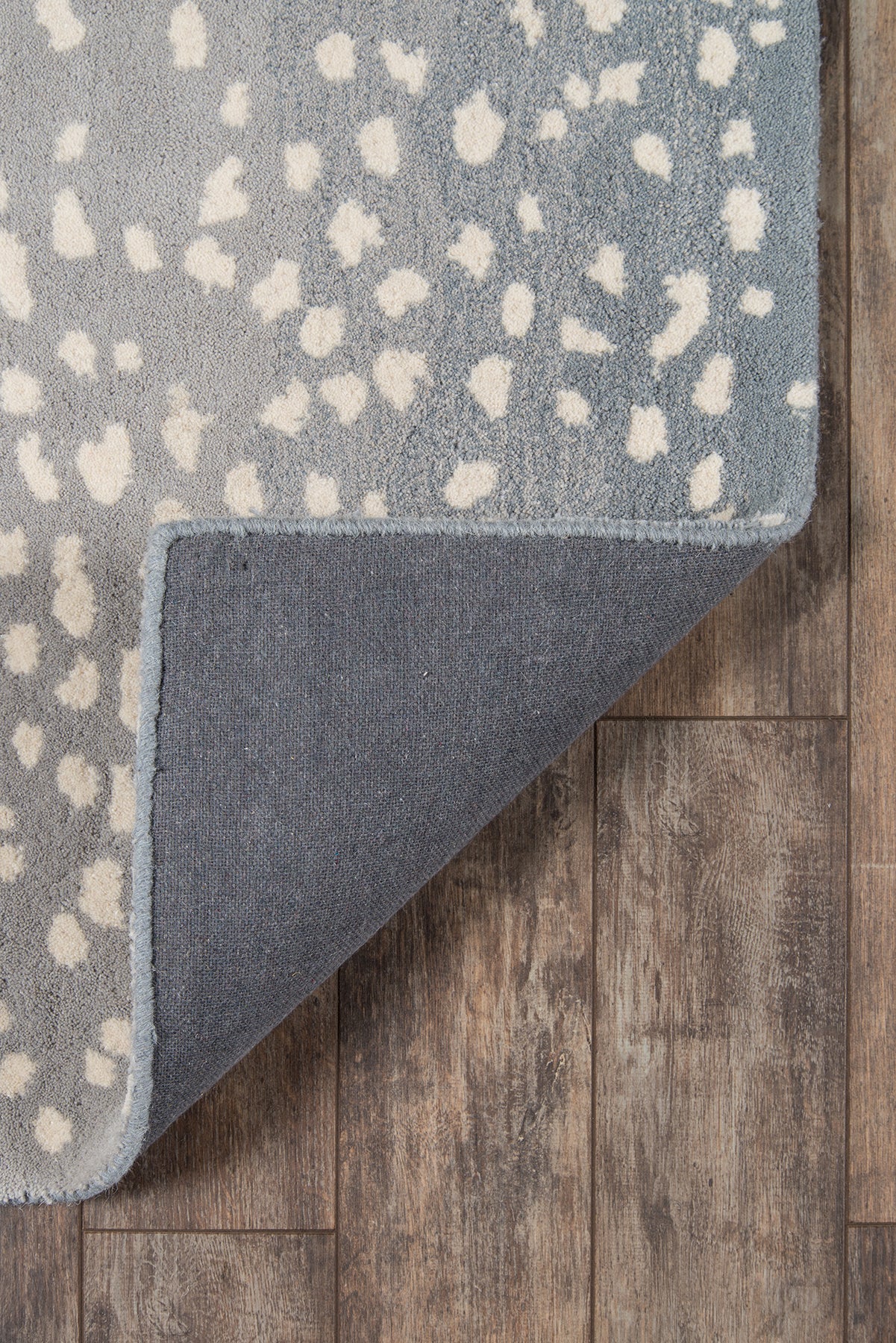 Blue-gray rug with irregular white specks; corner folded up to reveal solid blue backing on wood floor.