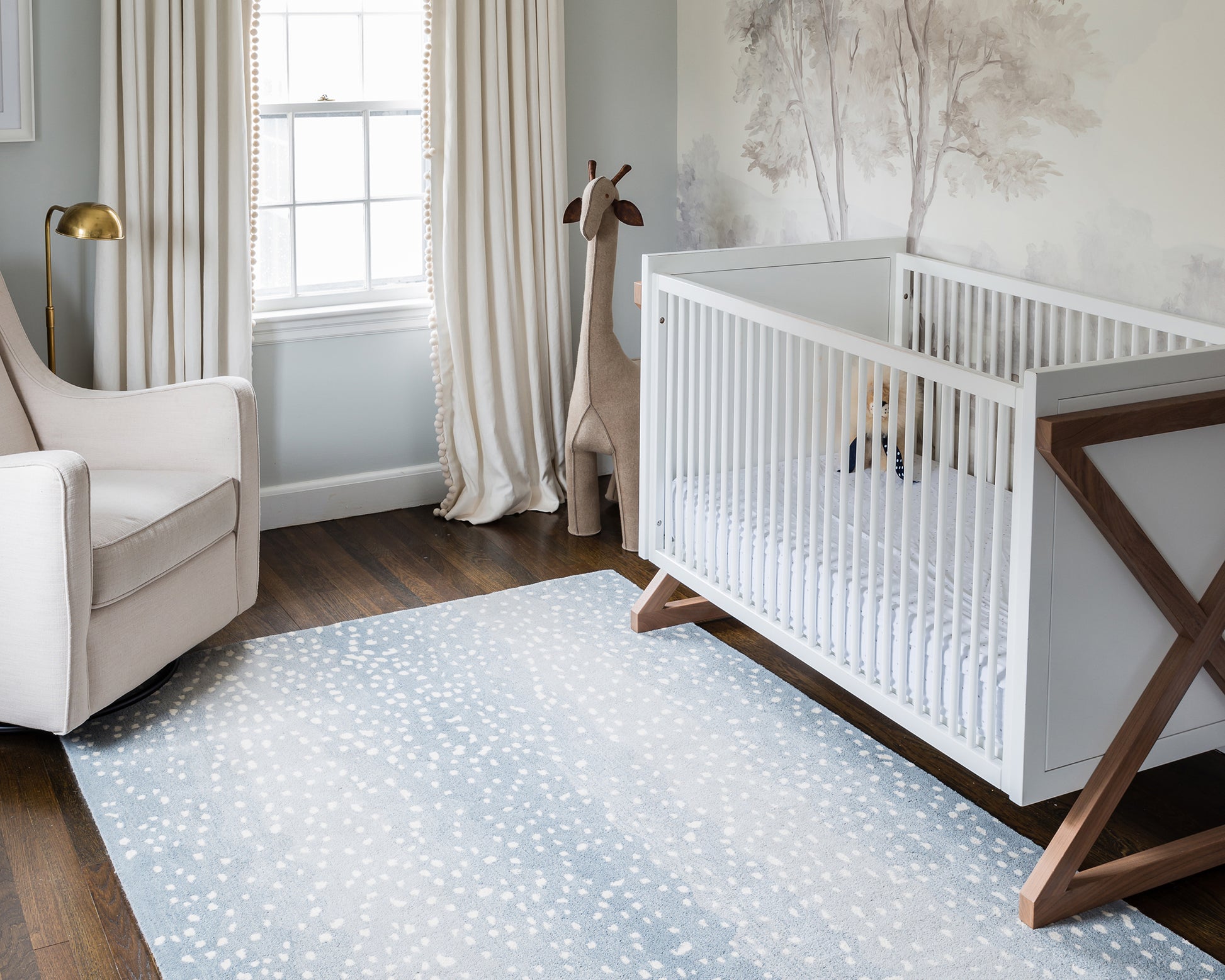 White wooden baby crib with slatted sides and angled legs; modern nursery with a blue speckled rug and beige armchair.