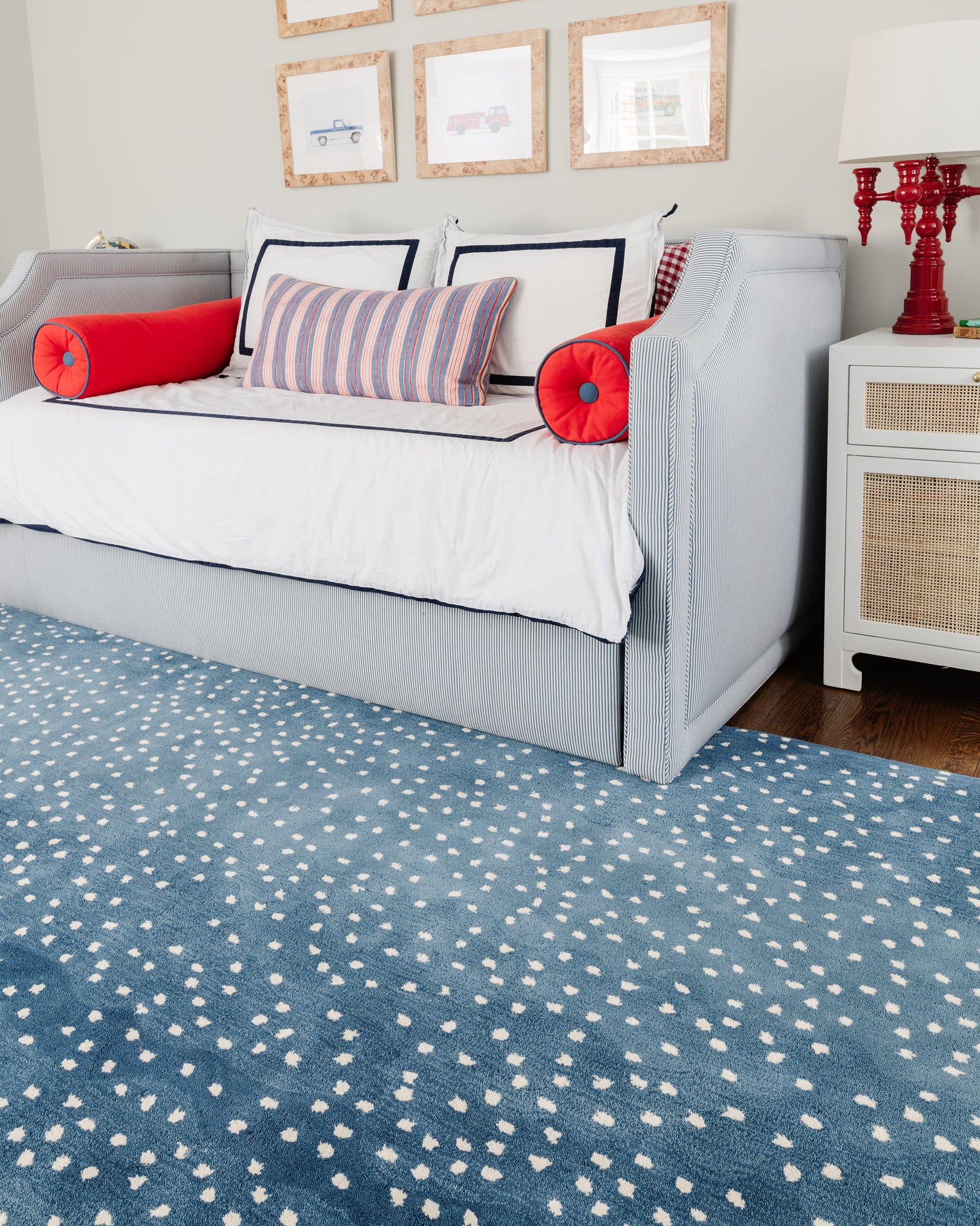 Light blue sofa with white cushions and red bolster pillows, blue dotted rug, white side table with red lamp in a cozy living room.