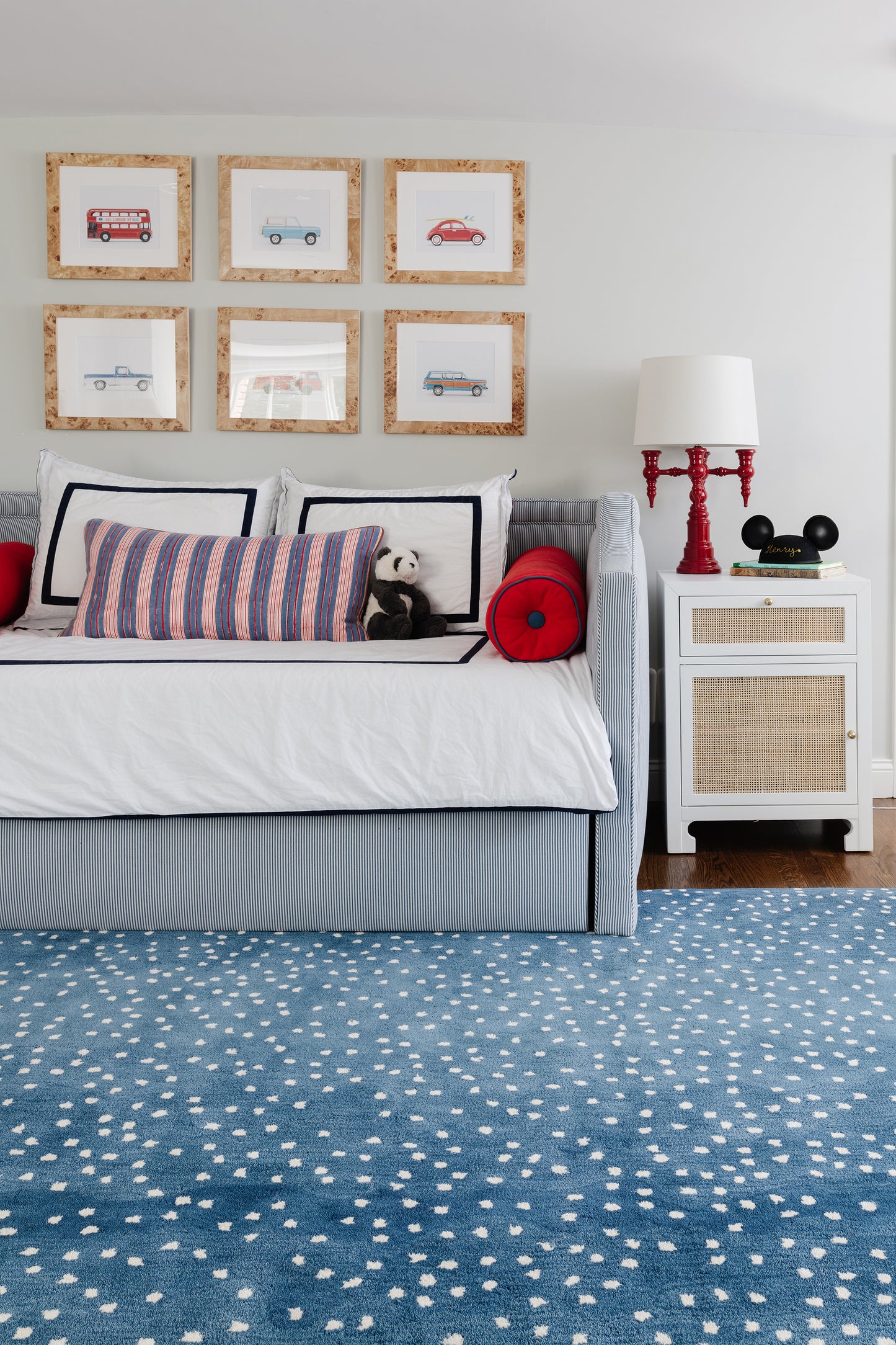 Coastal bedroom with blue striped bed, white bedding, cork-framed car prints, white nightstand, red lamp, blue dotted rug.