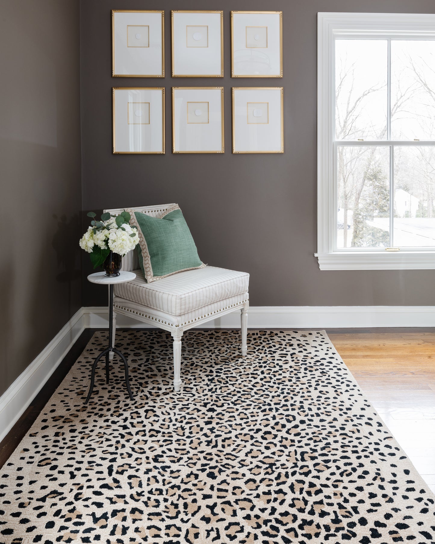Dark taupe wall, six gold-framed prints, white upholstered chair with green cushion, leopard rug, window.