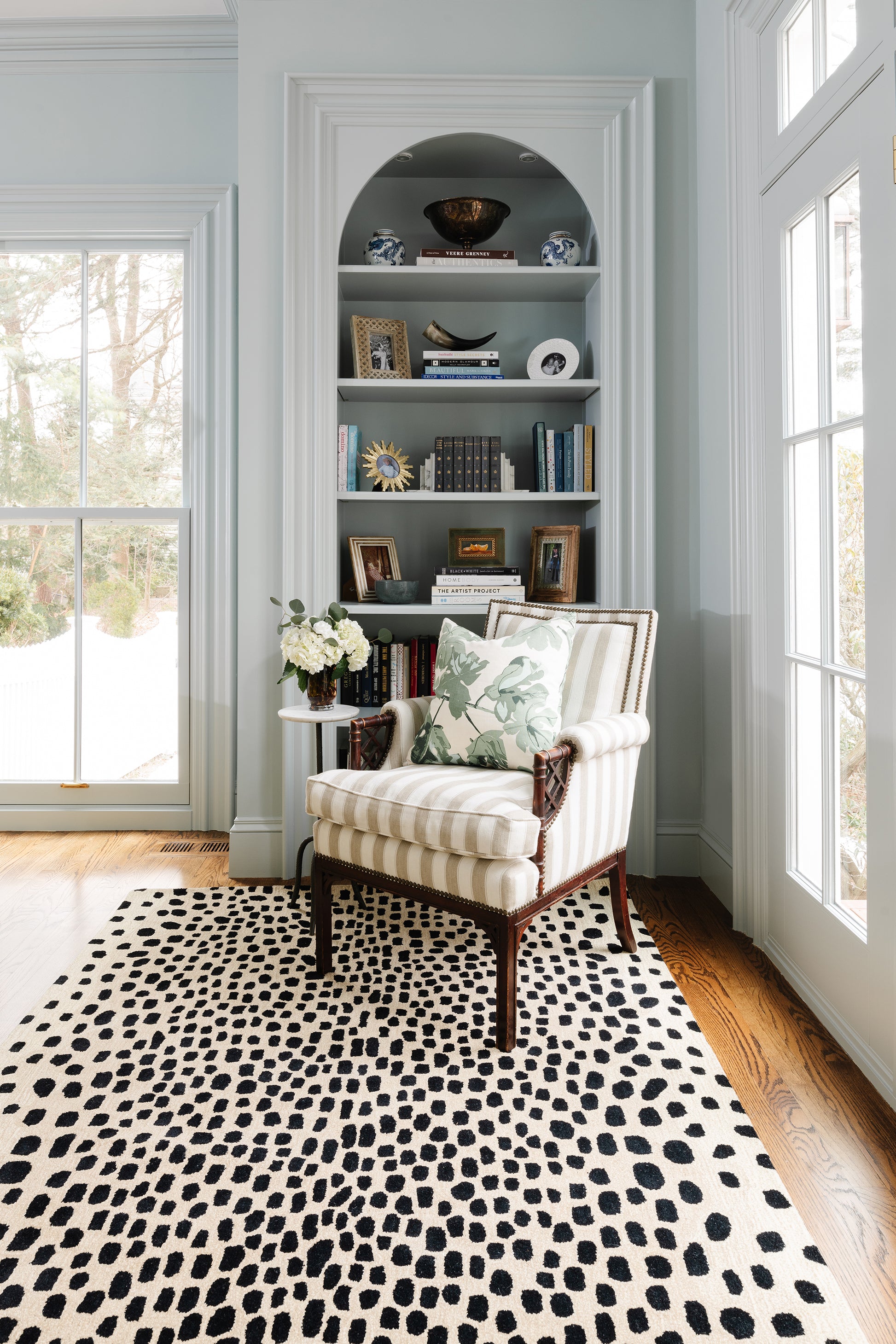 Light blue arched built-in bookshelf with a cream striped armchair, vase of white flowers, and a black-and-white spotted rug.