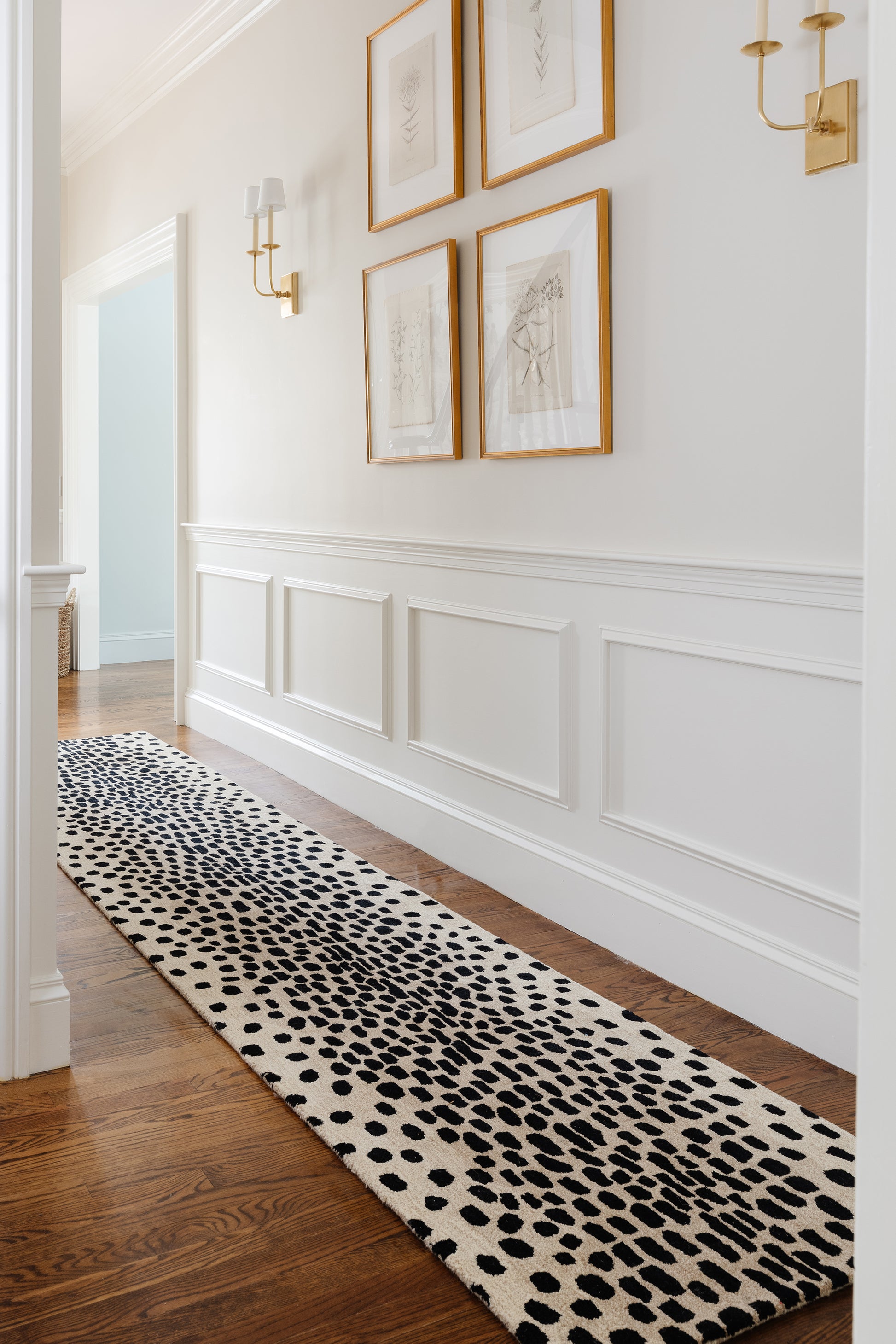 Long black and white spotted runner rug along a white wainscoted hallway with framed botanical art and gold sconces.
