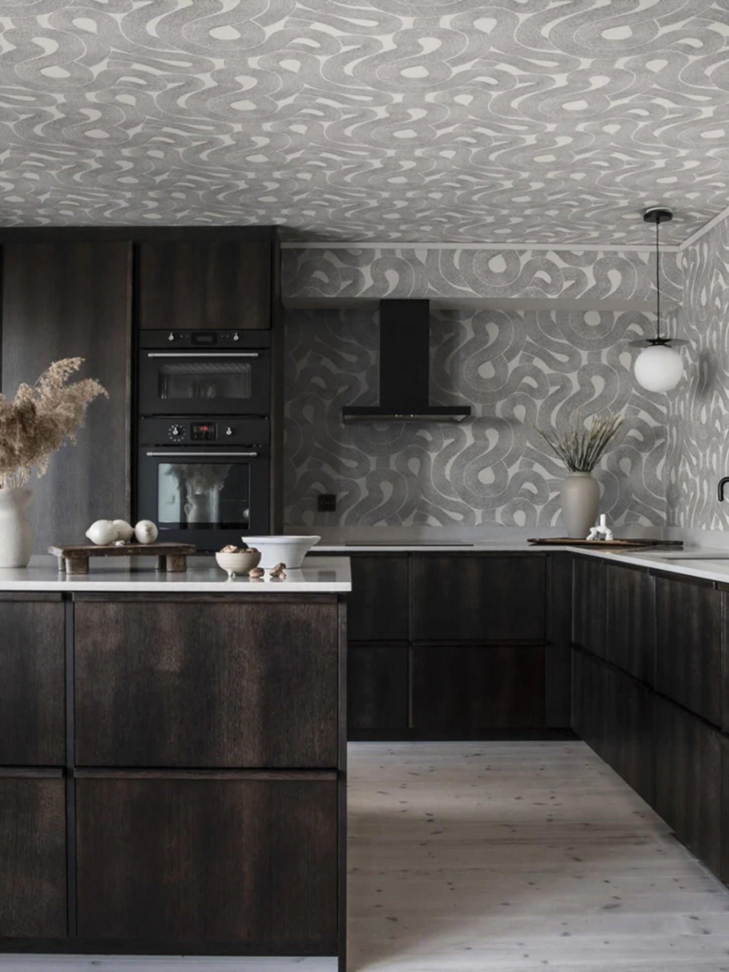 Modern dark wood kitchen with white marble island, built-in ovens, black range hood, and grey patterned wallpaper.