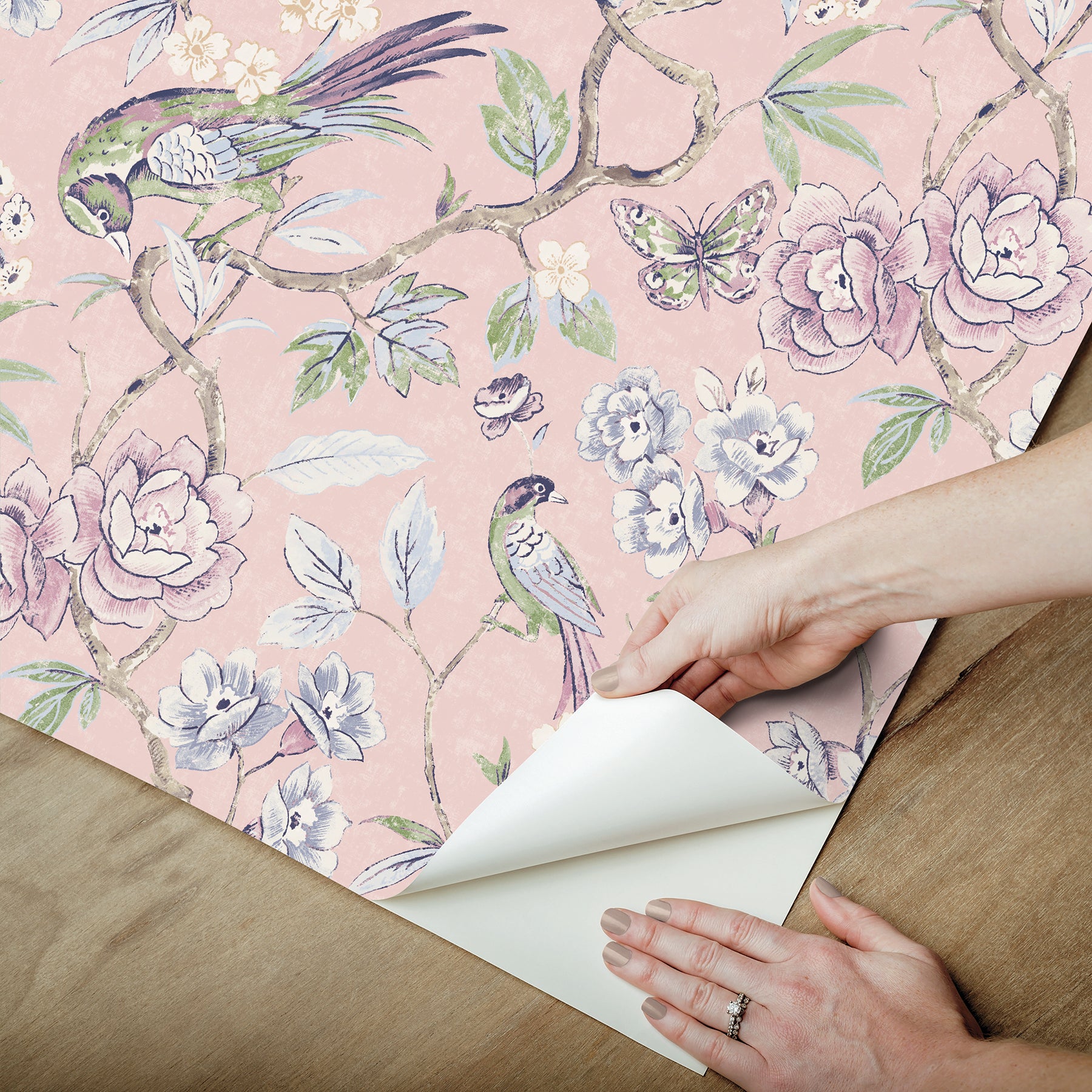 Pink floral wallpaper with birds and blossoms; hands peel back the sheet to reveal white backing.