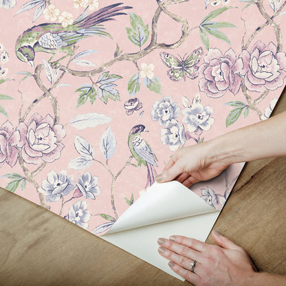 Pink floral wallpaper with birds and blossoms; hands peel back the sheet to reveal white backing.