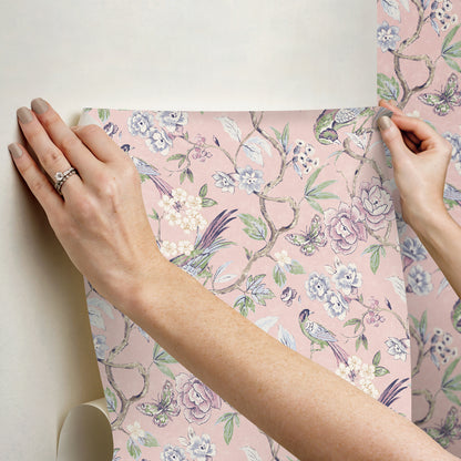 Hands hold a pink floral wallpaper swatch with lavender, white blossoms & green leaves on a soft pink background.