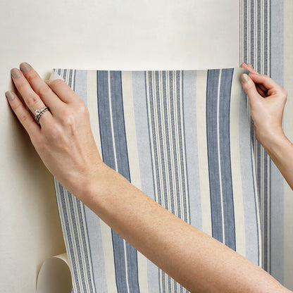 Hands smoothing blue and white striped wallpaper on a wall, roll visible in corner; coastal pattern.