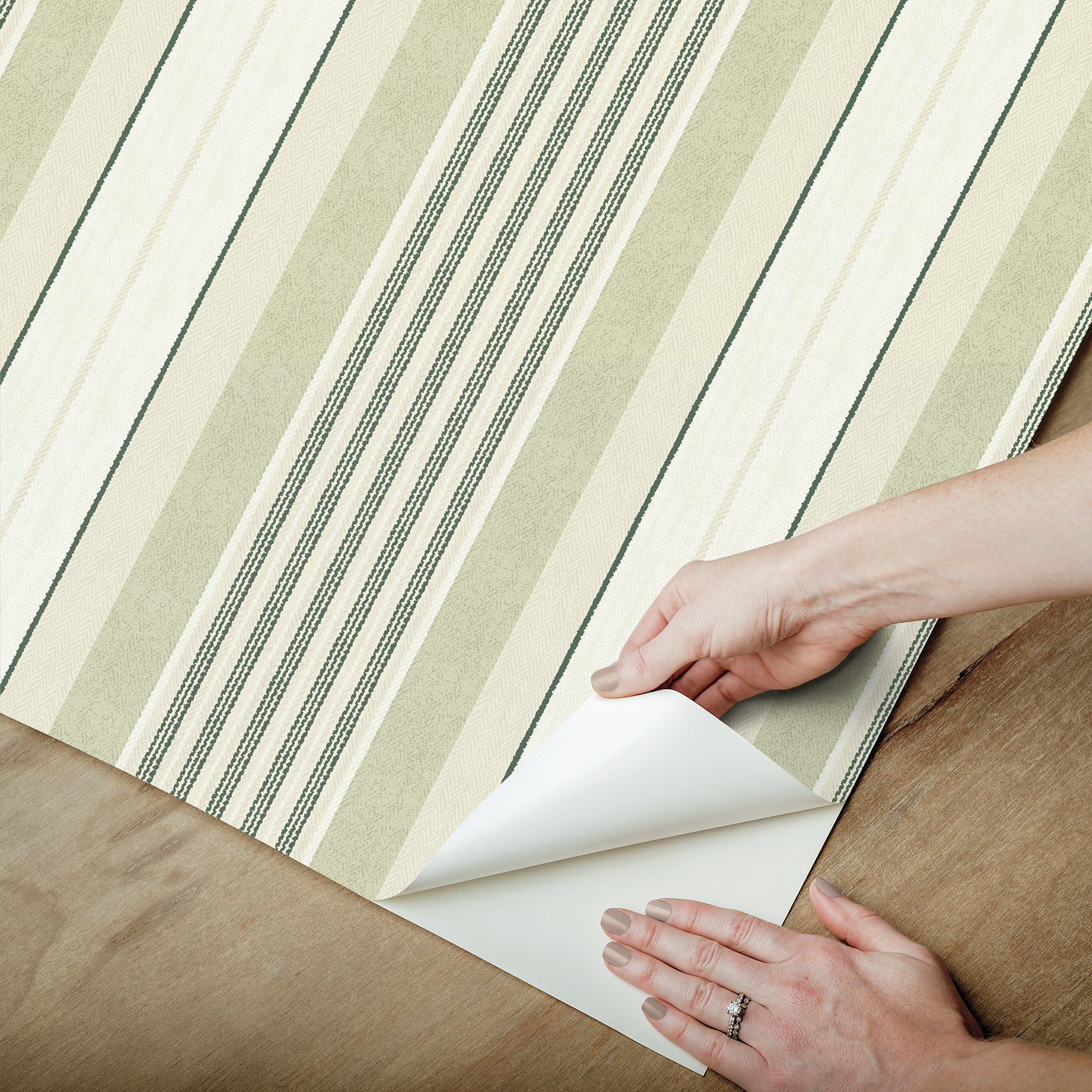Green striped wallpaper being peeled back on a wooden floor; curling corner, hand with ring.