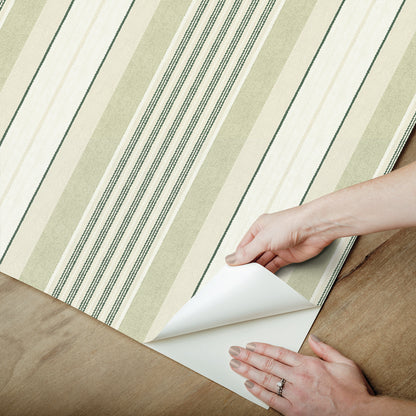 Green striped wallpaper being peeled back on a wooden floor; curling corner, hand with ring.