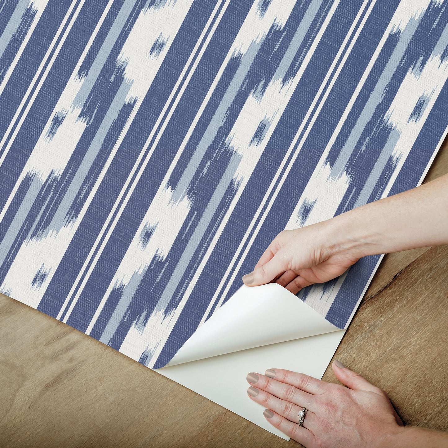 Blue and white ikat-striped wallpaper being peeled back on a wooden floor, hands pulling the corner to reveal white backing.