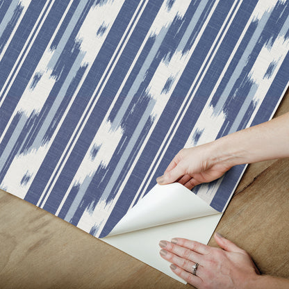 Blue and white ikat-striped wallpaper being peeled back on a wooden floor, hands pulling the corner to reveal white backing.