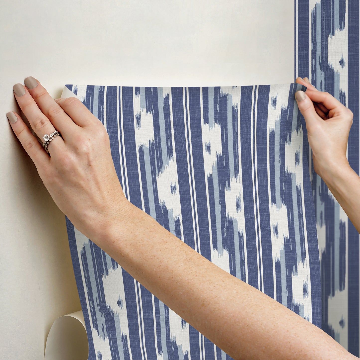 Hands smoothing a blue and white ikat-style wallpaper sheet onto a wall.