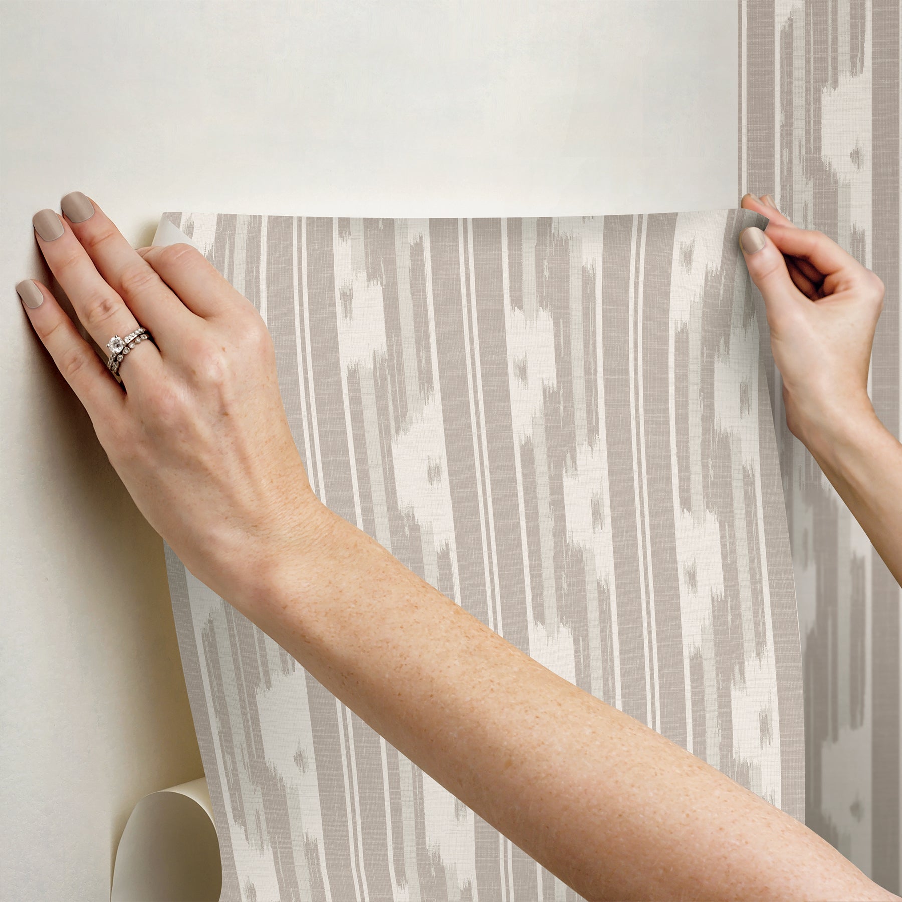 Hands smooth gray and white vertical striped wallpaper being applied to wall.