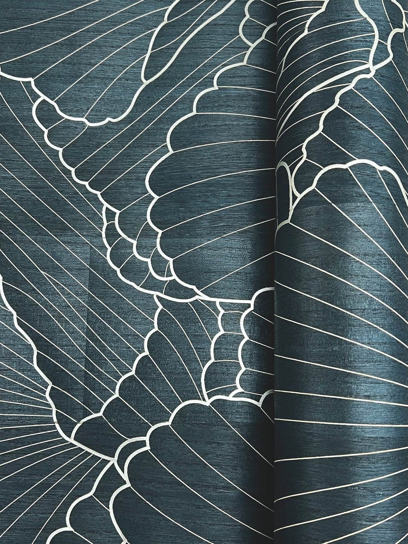 Blue linen-like fabric with white botanical outline pattern of large leaves, rolled at the right edge, close-up detail.