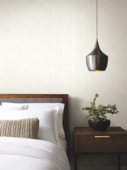 Bedroom wall covered with herringbone wood veneer Apple Blossom wallpaper; white bedding, dark wood headboard and nightstand, beige knitted cushion, black pendant lamp, and a potted plant.