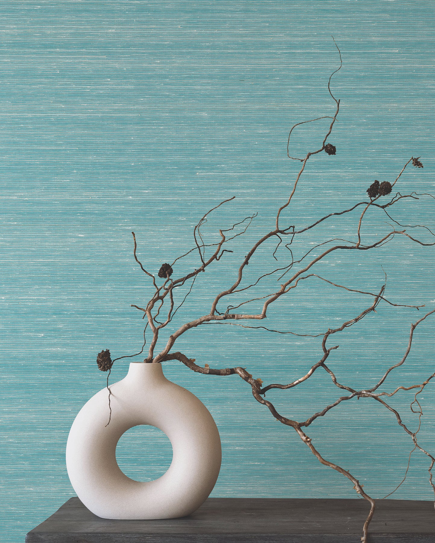 Room scene with turquoise textured wallpaper, a dark wood shelf, and a white donut-shaped ceramic vase with bare twigs and seed pods.
