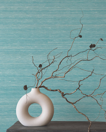 Room scene with turquoise textured wallpaper, a dark wood shelf, and a white donut-shaped ceramic vase with bare twigs and seed pods.