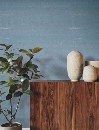 Blue textured wall, dark wood cabinet, tall beige ceramic vase and smaller vases, with a leafy plant on the left.