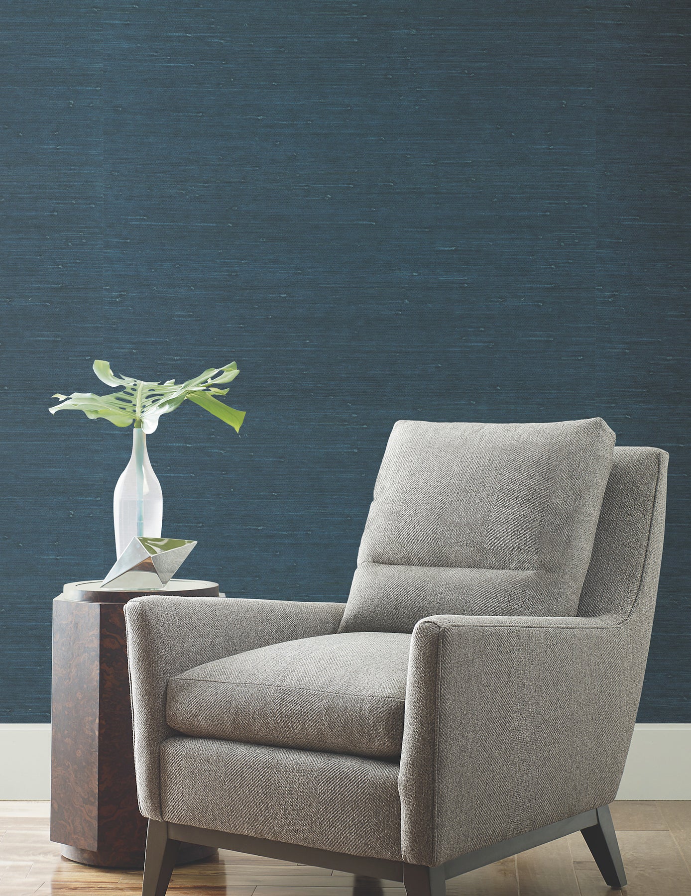Grey fabric armchair with dark wooden legs beside a round wooden side table and white vase with plant on a blue textured wall.