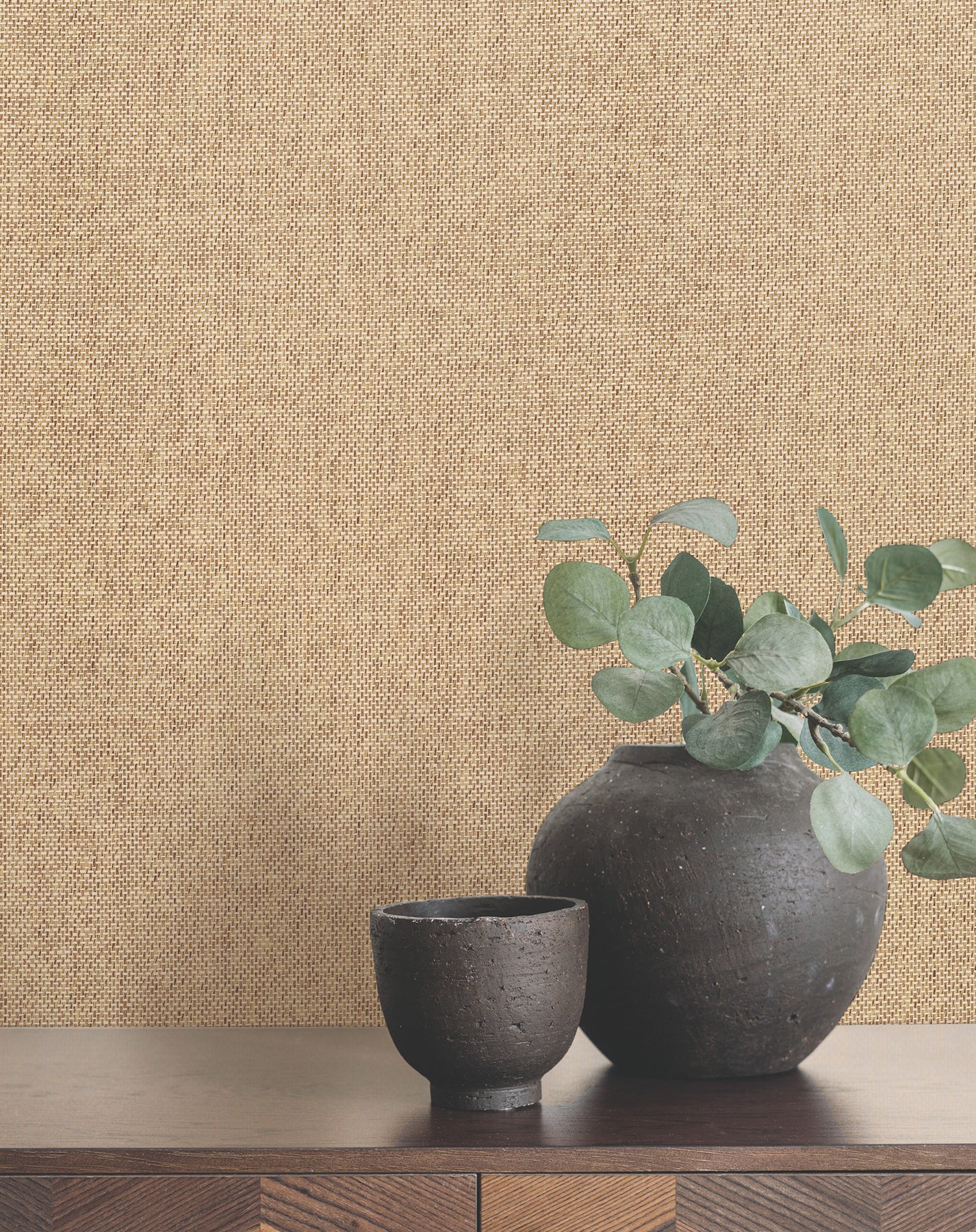 Butter paperweave wallpaper with a subtle woven texture; in front, a pair of matte black ceramic vases with eucalyptus on a wooden shelf.