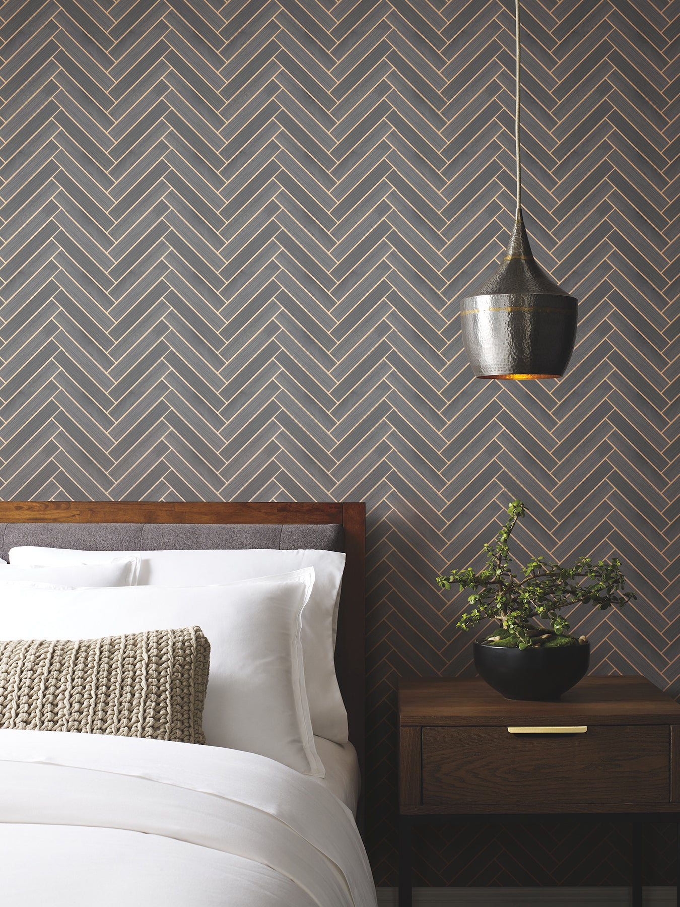 Bedroom with herringbone wallpaper, wooden bed, white linens, beige knit pillow, black pendant lamp, plant on nightstand.