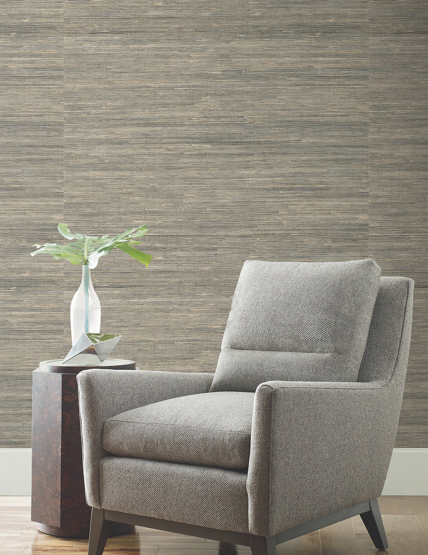 Grey fabric armchair with rectangular cushions and dark wooden legs, beside a small table with a ceramic vase and plant.