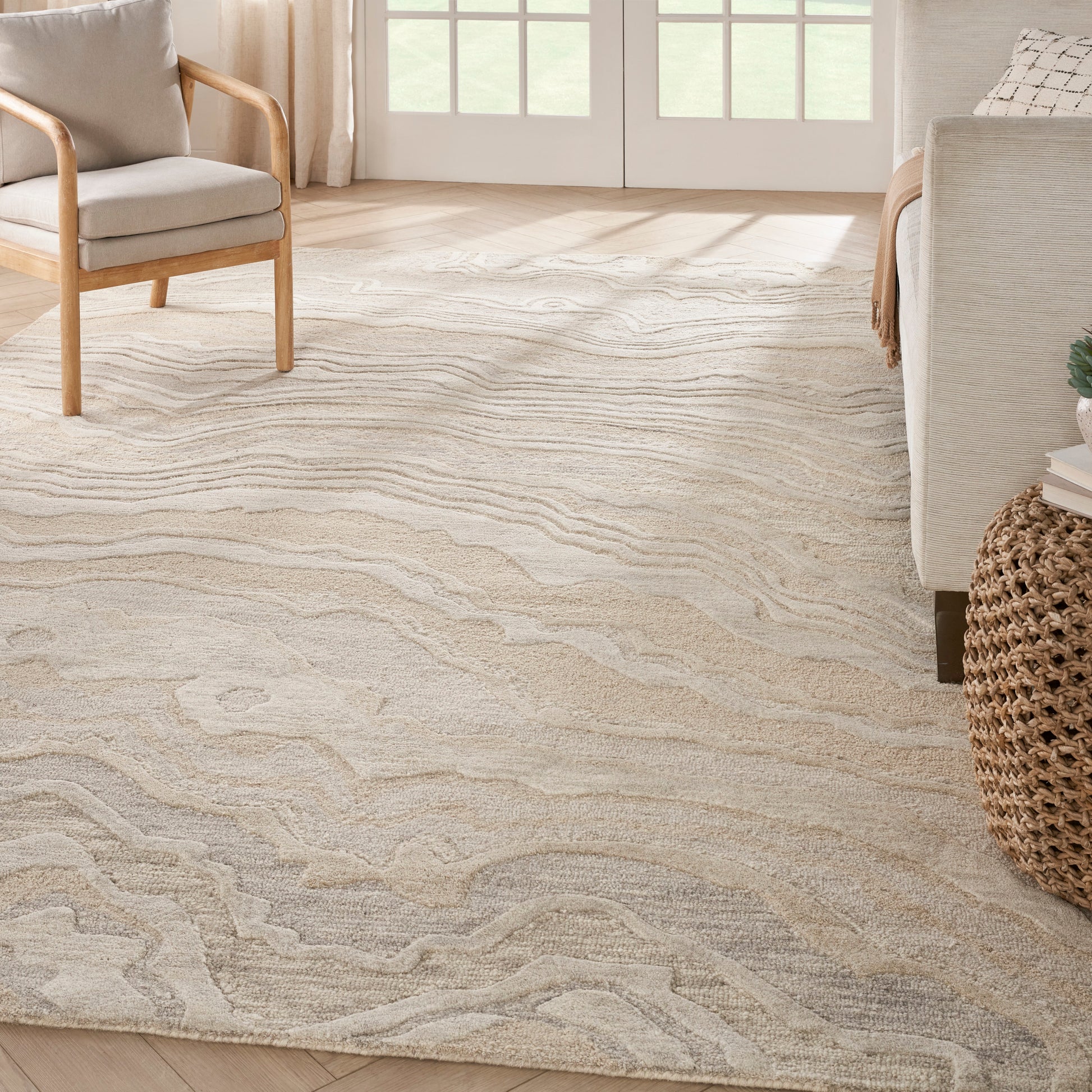 Neutral beige area rug with pale gray wave pattern in a bright living room with light wood furniture and a wicker accent.