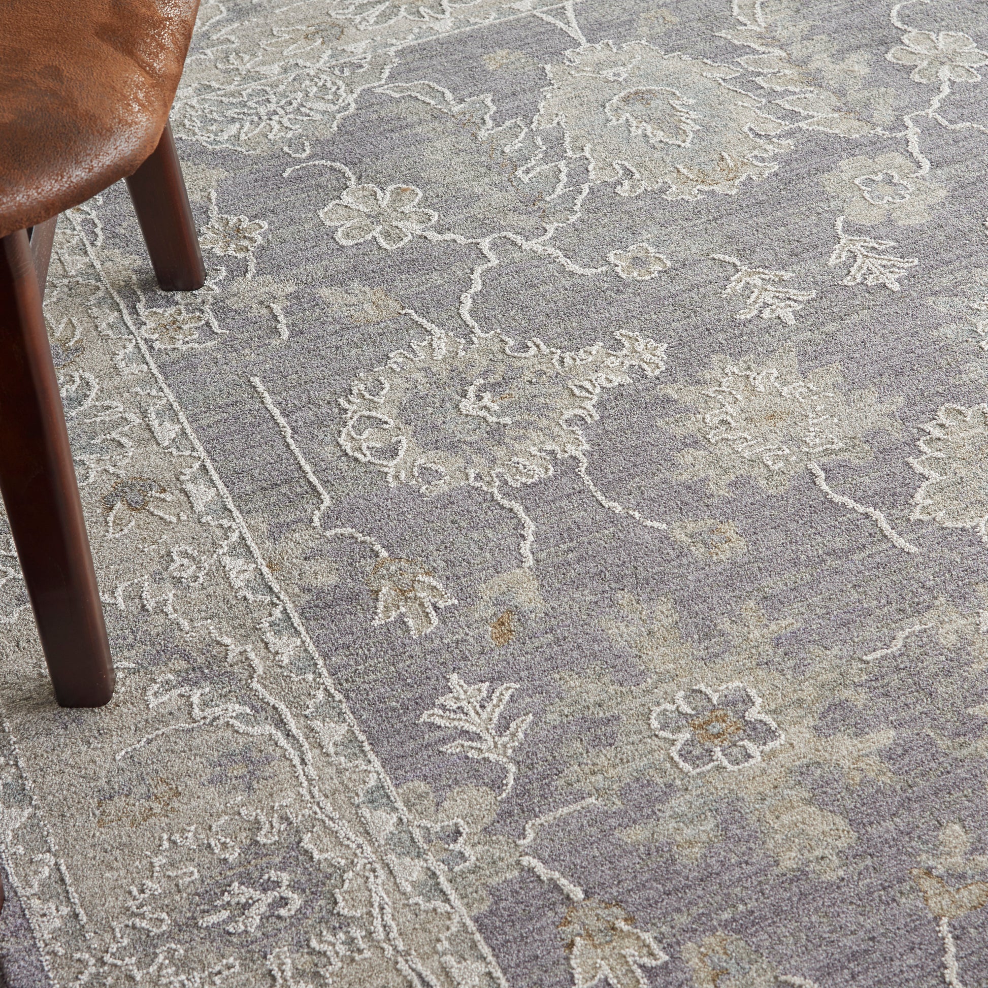 Close-up of a muted purple-gray floral rug with ivory outlines; wooden chair leg visible on the left.