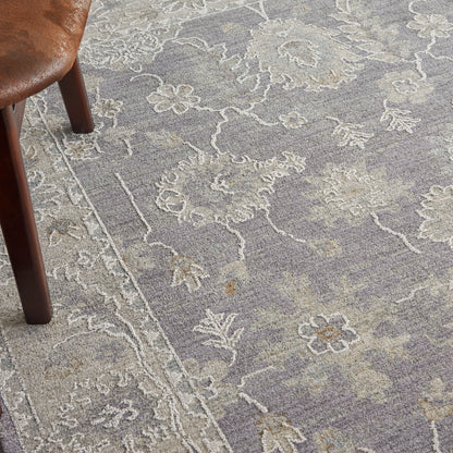 Close-up of a muted purple-gray floral rug with ivory outlines; wooden chair leg visible on the left.