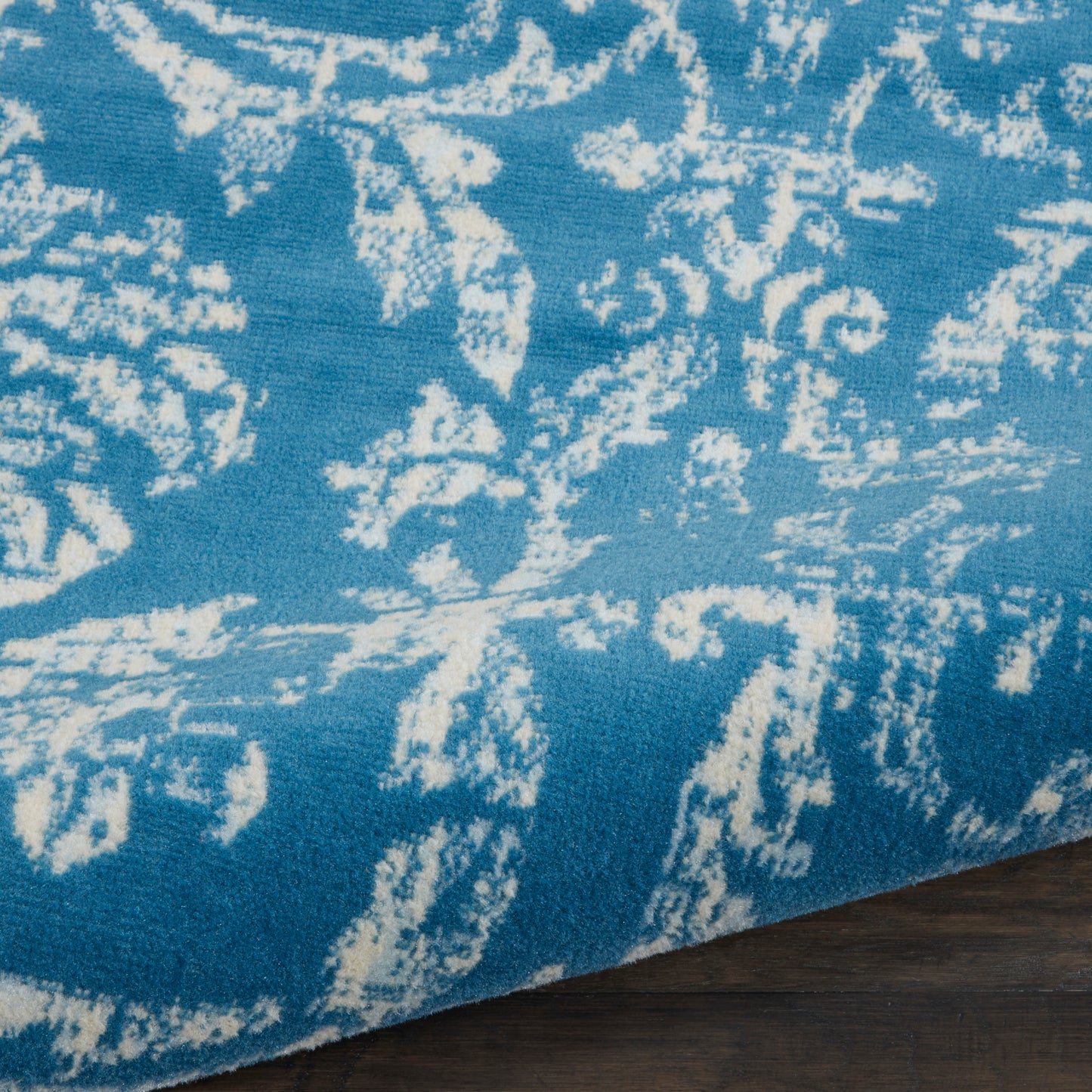 Blue rug with white abstract floral motifs, edge folded over dark wooden floor.
