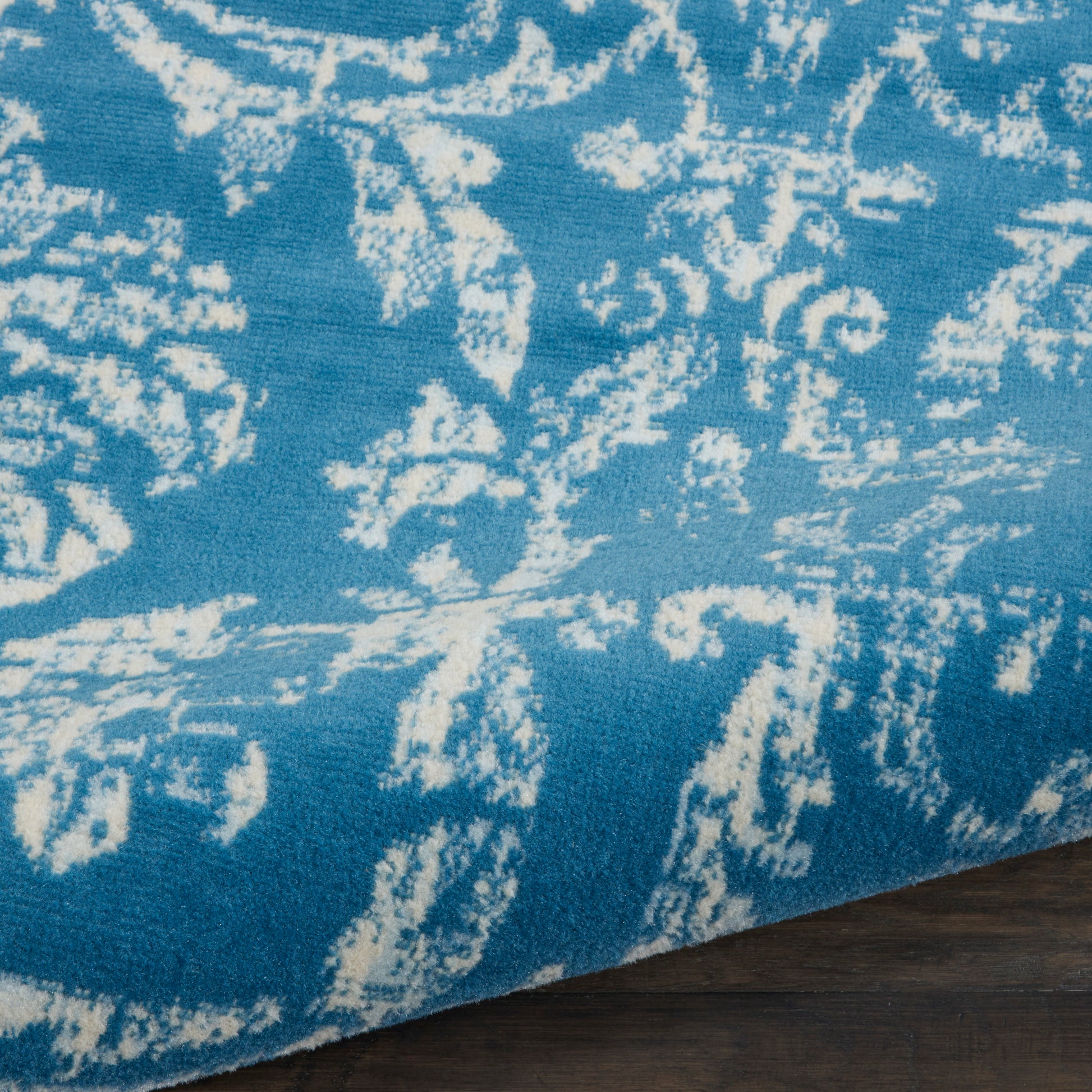 Blue rug with white abstract floral motifs, edge folded over dark wooden floor.