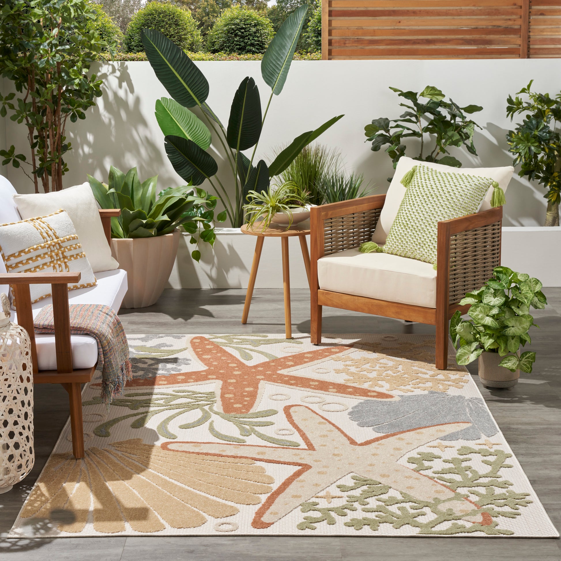 Outdoor patio with wooden frame chairs, white cushions, green pillows, a round table, lush plants, and a botanical beige rug.