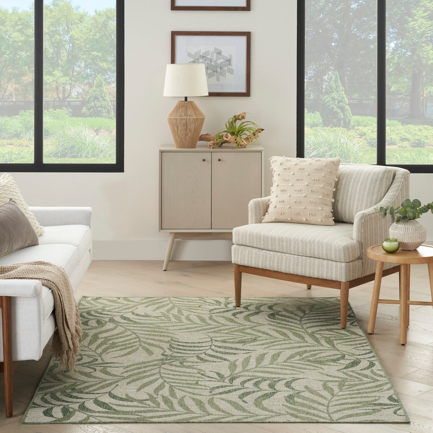 Neutral living room with cream sofa and striped armchair, green palm-pattern rug, wood cabinet, plant, and large windows.