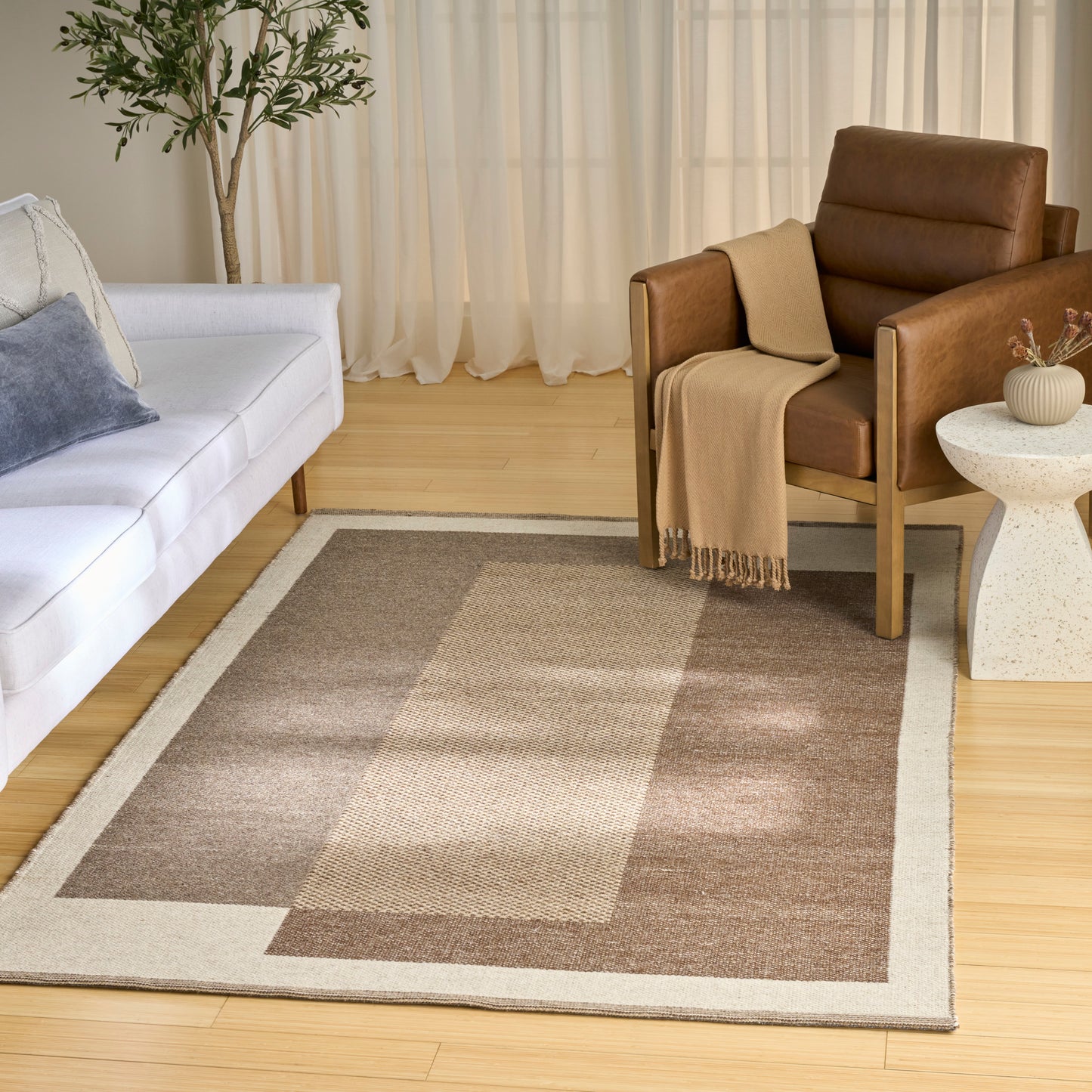Geometric beige-taupe area rug with border in a modern living room; white sofa, brown leather chair, and sheer curtains.