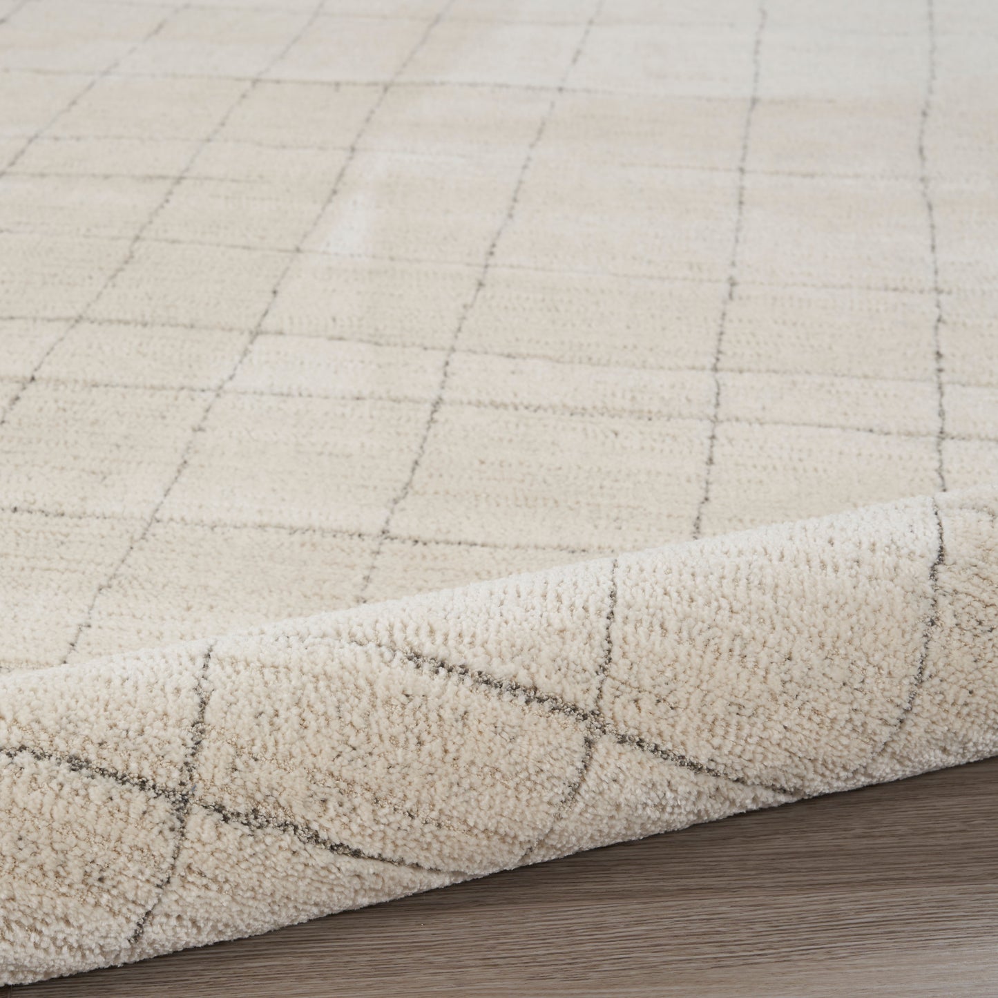 Beige area rug with a subtle gray grid pattern and plush texture, rolled corner on a wood floor.