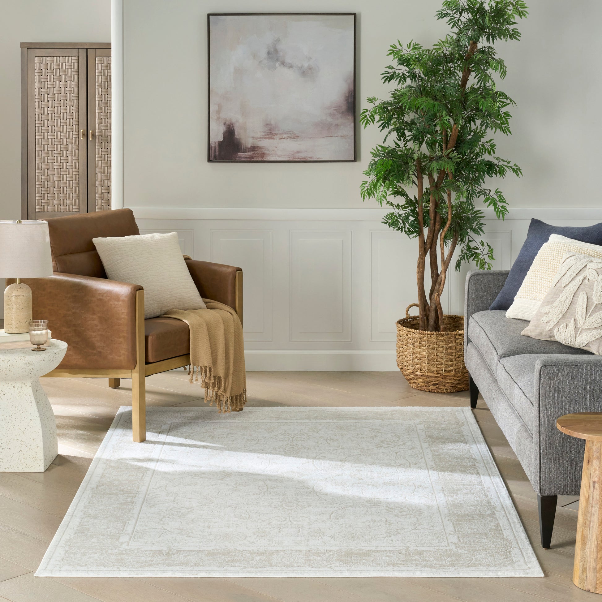 Neutral area rug with subtle pattern in a light living room; gray sofa, brown leather chair, plant in woven basket, and abstract art.